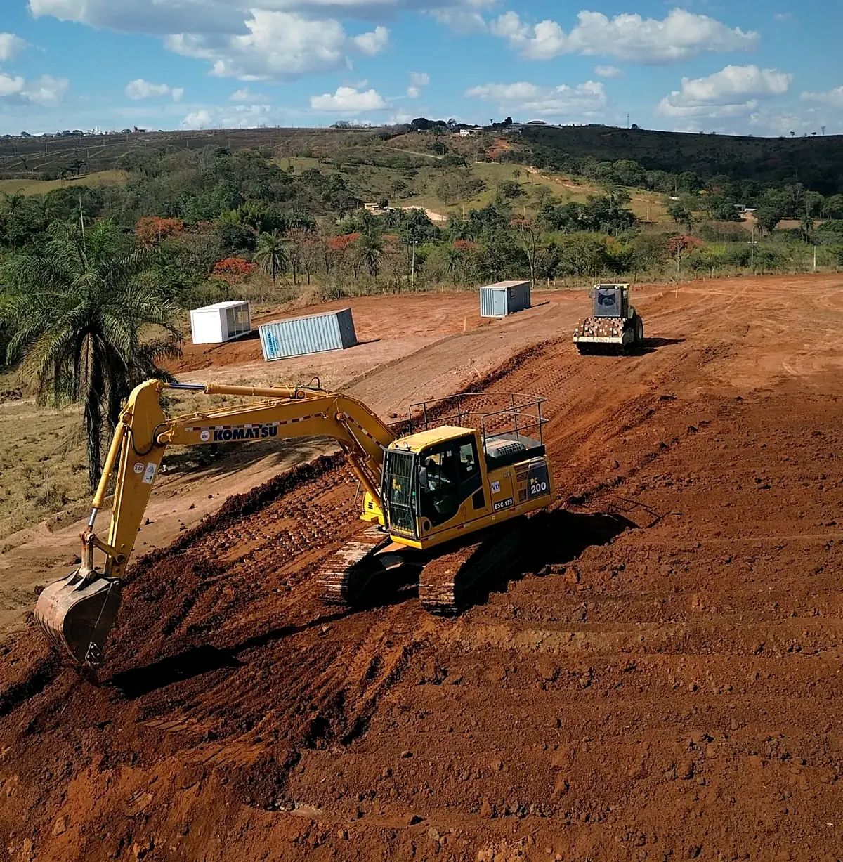 Escavadeira hidráulica Komatsu PC200 da TTL executando terraplenagem em talude de solo vermelho, com rolo compactador ao fundo, em Minas Gerais